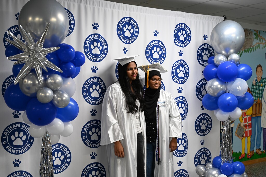 Two graduating seniors pose in front of the Latham Ridge backdrop adorned with balloon columns.
