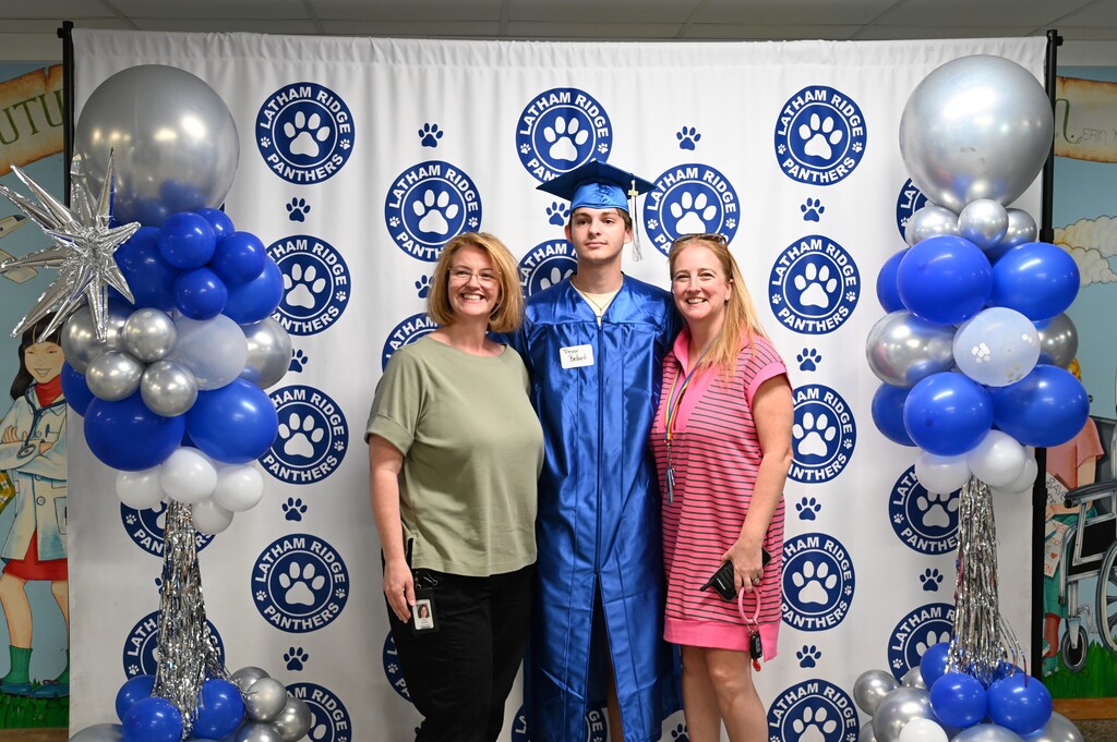 Teachers pose with a former student in front of a backdrop with balloons.