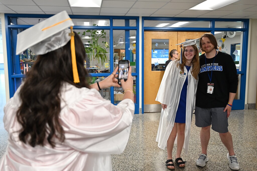 A student takes a photo on her phone of another senior and a former teacher.