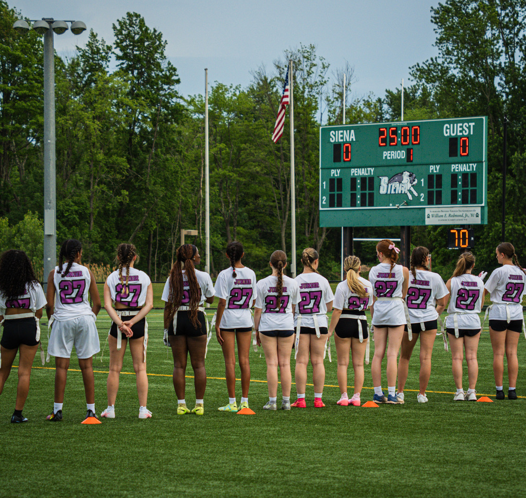 Juniors stand on the sideline looking at the American flag and the scoreboard.