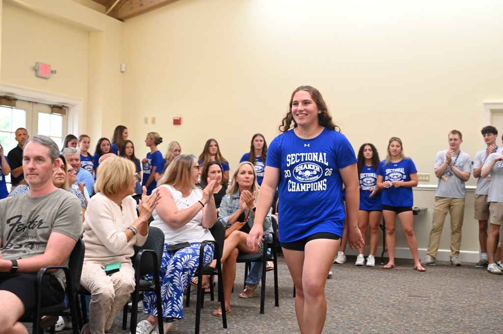 A member of the Shaker girls lacrosse team walks to the front of the room surrounded by family and friends seated in chairs.
