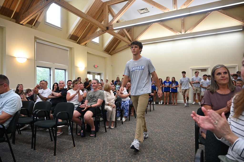A member of the Shaker boys lacrosse team walks to the front of the room surrounded by family and friends seated in chairs.