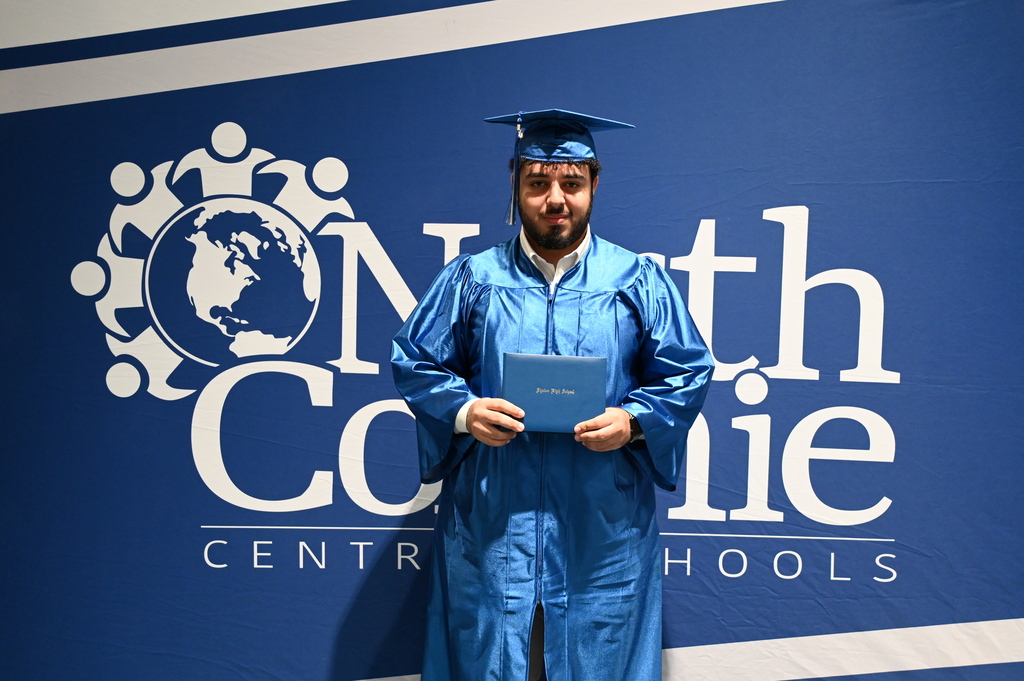 A student holds his high school diploma in cap and gown as he stands in front of a backdrop.
