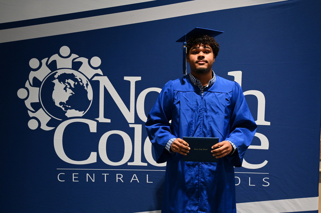 A graduate in cap and gown holds his diploma in front of a backdrop.