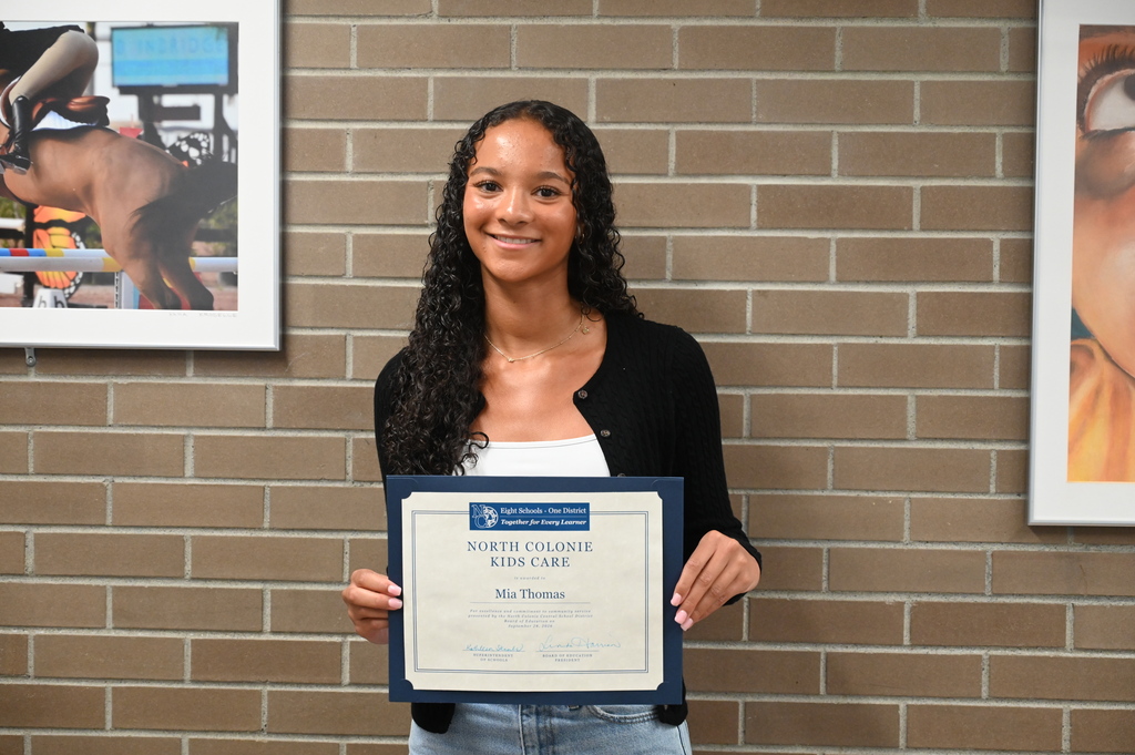 A student stands against a brick wall holding her certificate for Kids Care recognition.
