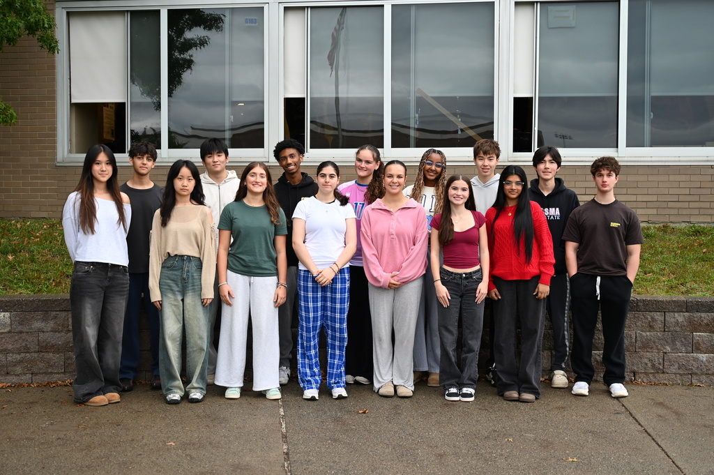 15 students pose in front of a concrete wall, being recognized for being selected to the NYSSMA All-State Festival.