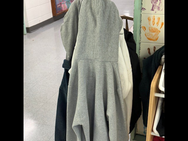 A close-up of a gray jacket hanging on a hook among other jackets. The floor has white tiles.
