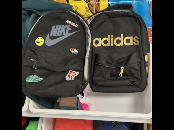 Two black backpacks with brand logos: Nike and Adidas. Each has zippers and pockets. Displayed on a white tray.