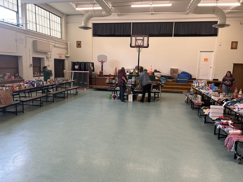 A few people gathered in a school gymnasium surrounded by clothes, toys, and food for a giveaway
