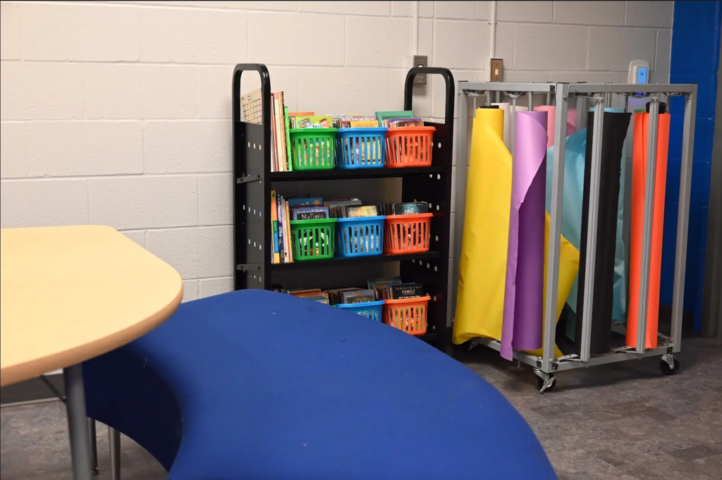 A table, bench, cart full of books, and rolls of paper in a classroom