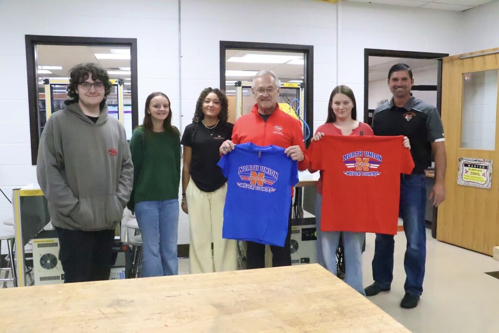 Five people stand behind a table, holding shirts. Two hold up red shirts and three hold up blue shirts.