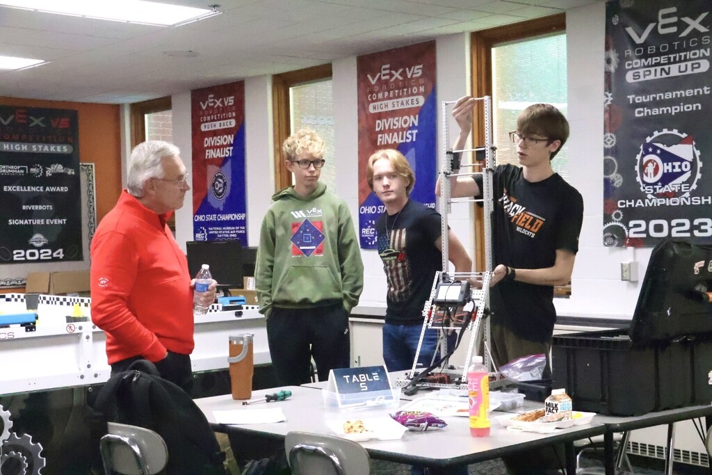 Four people stand around a table in a classroom. Two hold up a robotic arm. One stands on a chair.