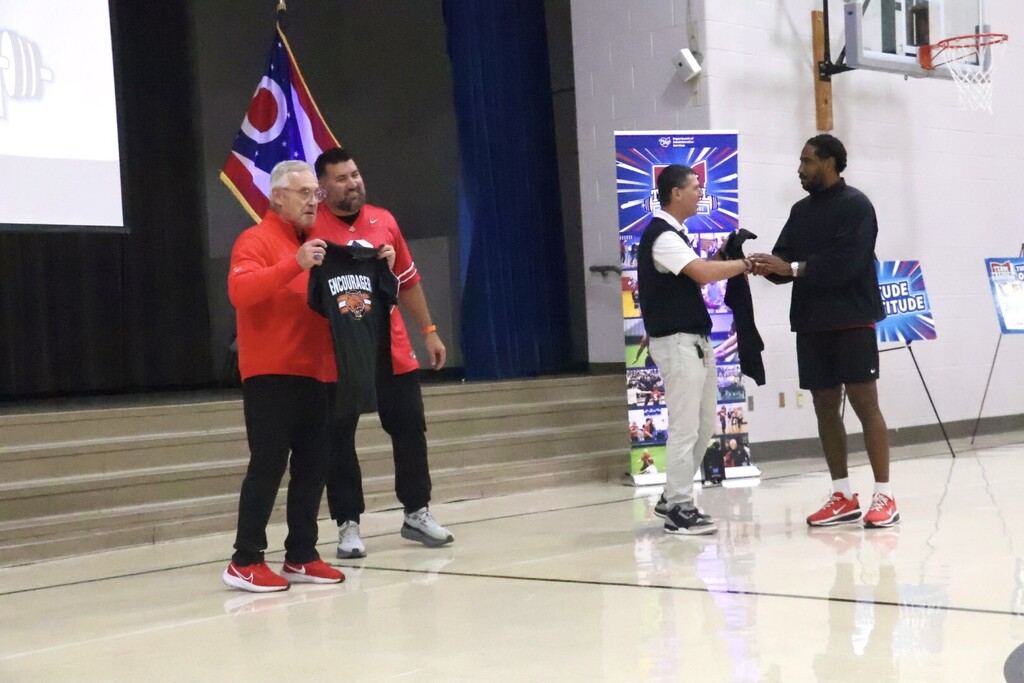 Four men on a basketball court. One man holds a jersey. Two others hold hands, shaking.