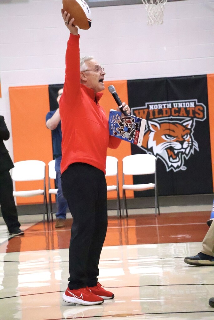 Man in red top holds a football and microphone in a gymnasium with chairs, and a mascot banner.