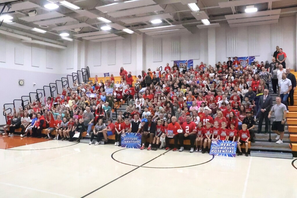 A large group in red shirts gathers in a gym with bleacher seating and a basketball court.