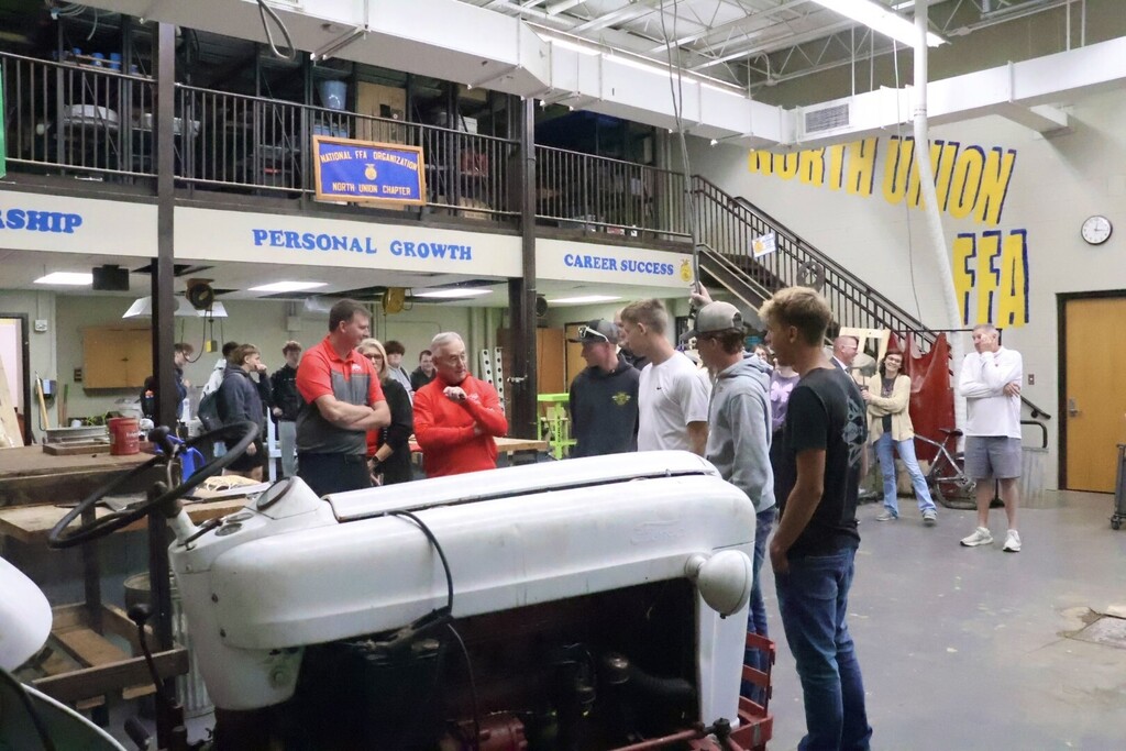 A group of people are gathered around a large, white machine in a large room. A banner hangs on the wall.