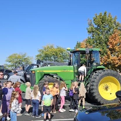 A group of children stands around a large green tractor with yellow wheels. Behind them, a person is climbing onto the tractor.