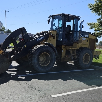 A yellow construction vehicle, possibly a John Deere loader, parked on a paved lot with a tree.
