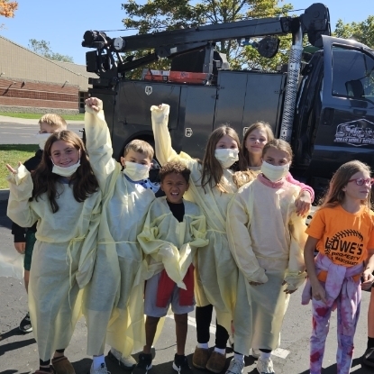 Group of children wearing masks and protective suits standing in front of a black tow truck.