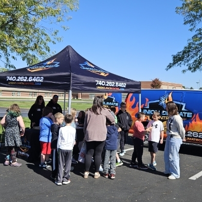 A group of children and adults gather under a tent with a fire graphic. They are looking at something on a table.