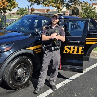 Police officer in uniform standing in front of a patrol car with the word "SHE" on the door.
