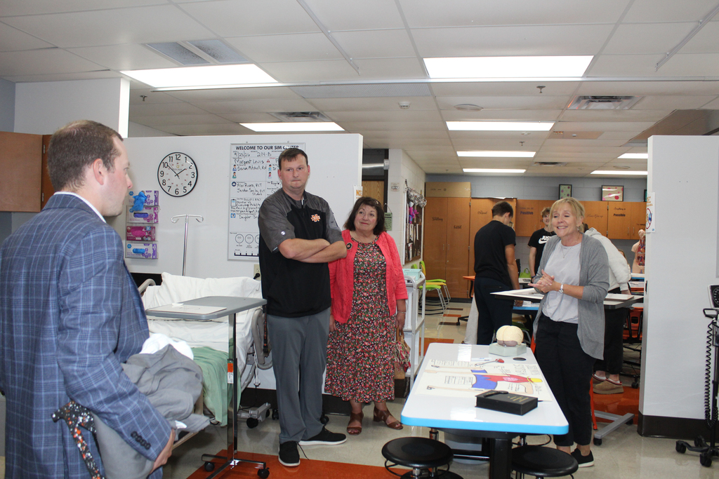 A group of people are in a hospital room. Three people are conversing while others are standing and observing.