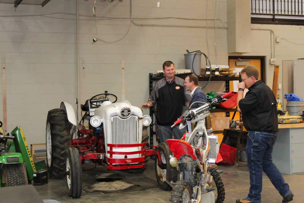 Three men in a garage with a red and white tractor and a red dirt bike. One man holds a camera.
