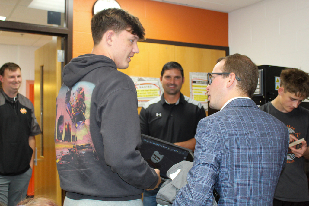 A group of men in a room. One man in a hoodie stands facing another in a suit.