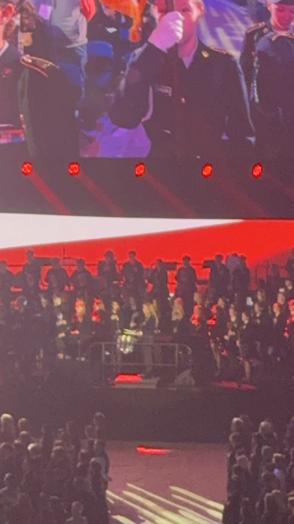 A large crowd in a venue with a stage, large screen, and red lighting; two people in military uniforms.