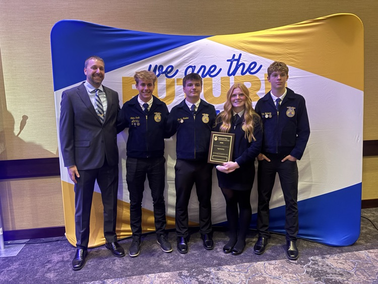 Five students and one adult stand behind a blue and yellow banner; the adult holds an award.