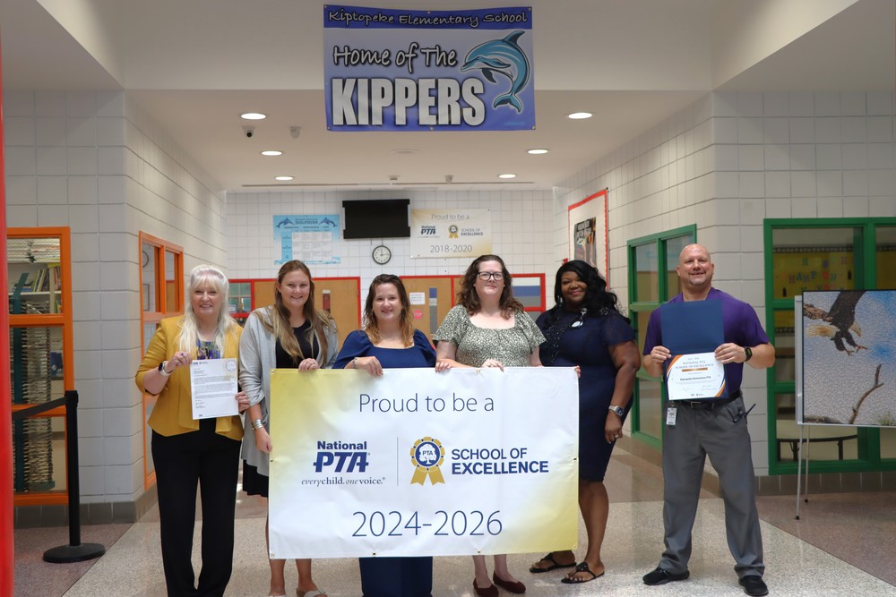 A photo shows five women on the left and one man on the right standing in a school hallway. They are holding a large gold and white banner that reads, "Proud to be a National PTA School of Excellence 2024-2026." The two individuals on the ends are also holding framed awards. A blue banner hanging in the hallway identifies the location as Kiptopeke Elementary School, Home of the Kippers.