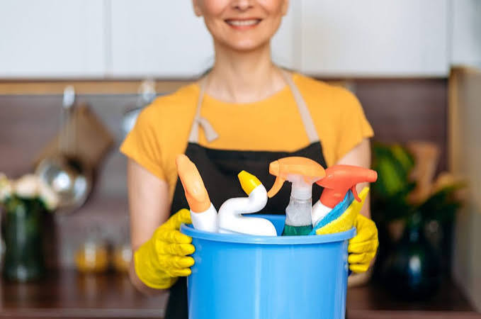 a woman in a yellow t-shirt is holding a blue plastic cleaning bucket with 4 bottles that are full of cleaning product