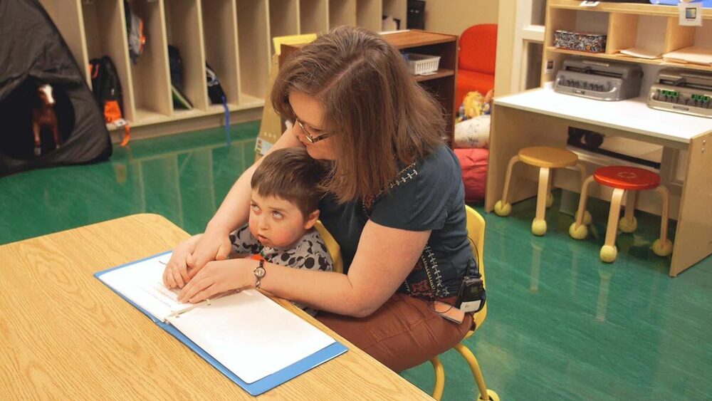 Teacher sitting behind student who is blind using hand under hand assistance