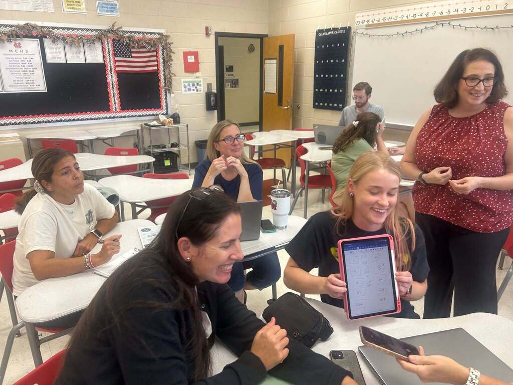 A classroom with desks, a teacher, and students. A teacher interacts with students using a tablet, and others engage with their laptops.