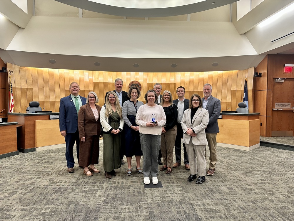 A group of people smiling to the camera with a woman standing in the front holding a small statute award. In the background is a city council chamber