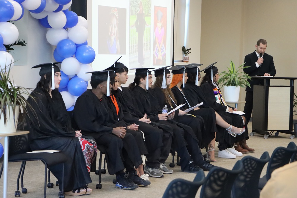 Graduates in their graduation gown and caps next to a man at a podium talking into to microphone