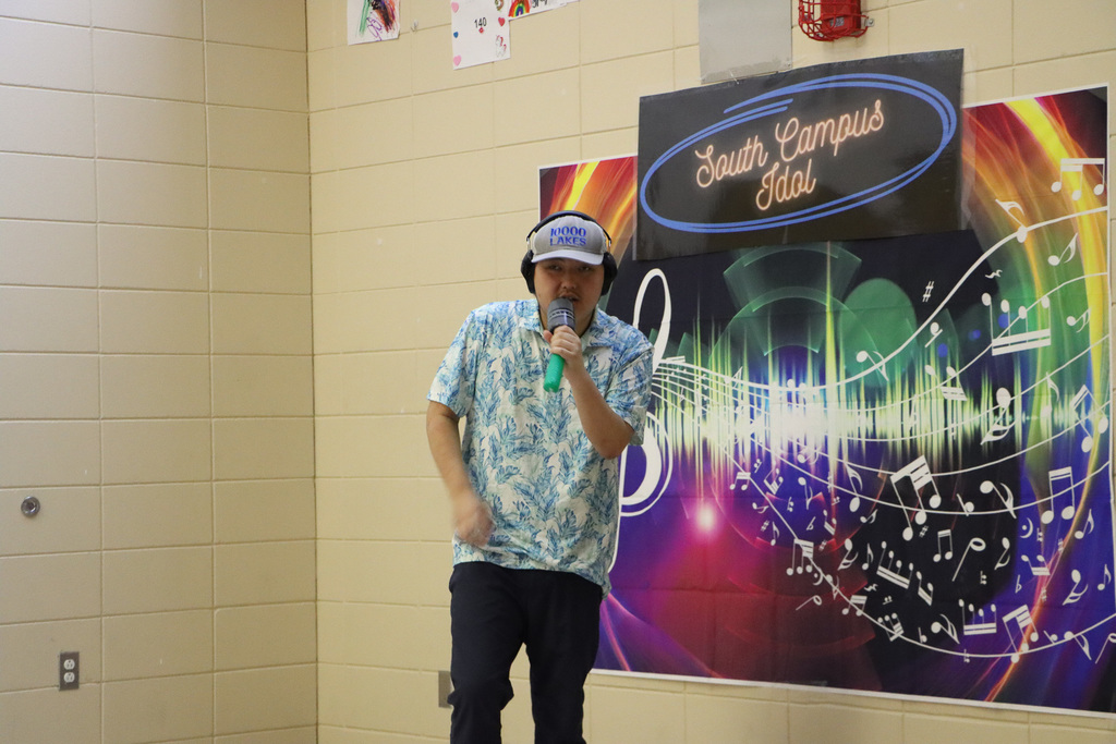 Image of a student singing into a microphone while standing in front of a background that has music notes and South Campus Idol.