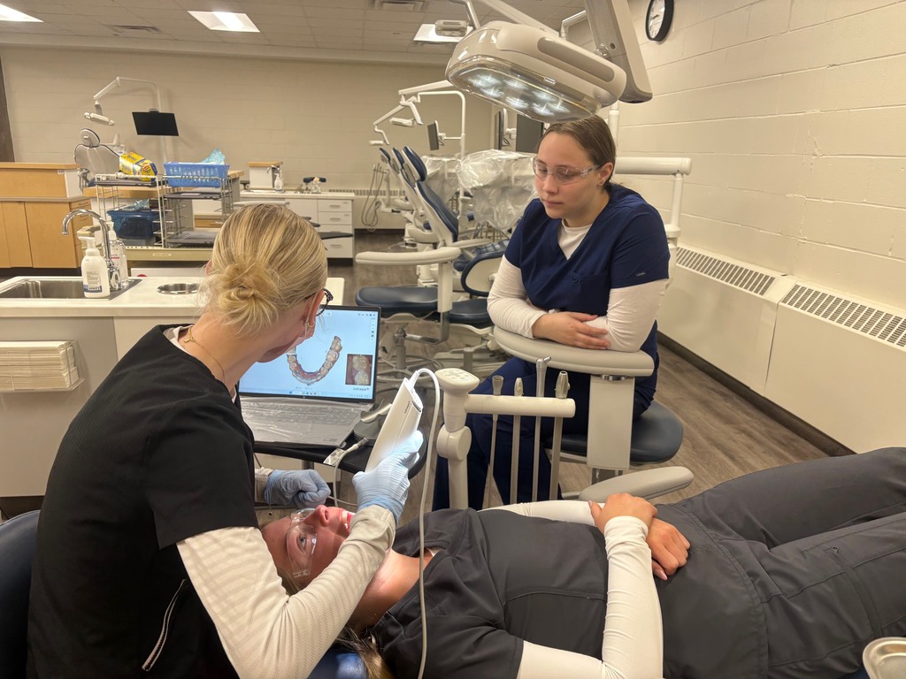 Student using a 3Shape TRIOS scanner to capture a 3D digital model/scan of classmate's teeth with another student observing them.