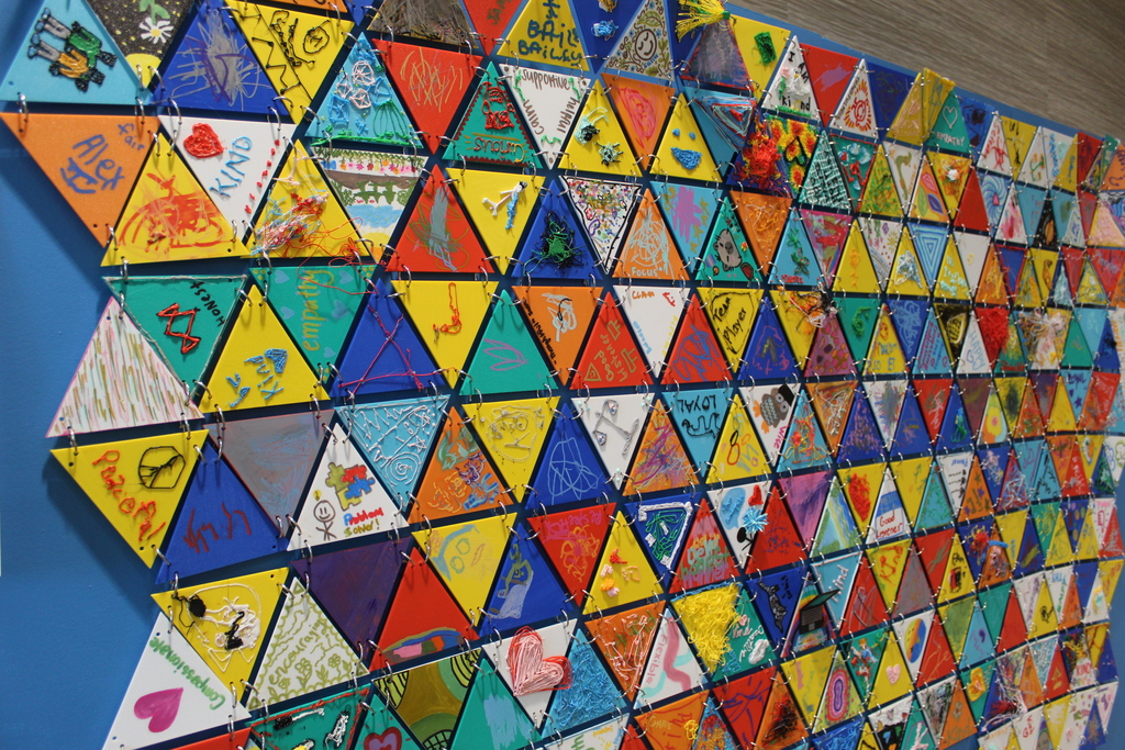 A wall of 3D printed triangles that are bound together by binder rings and each triangle has artwork by students and staff.