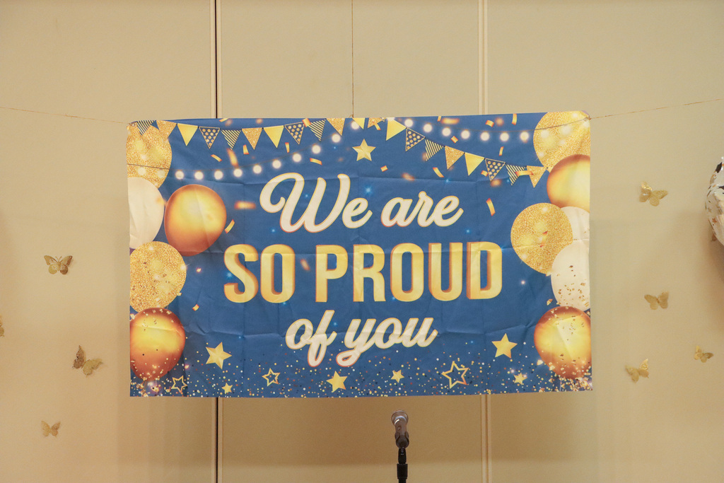 A blue banner that has gold balloons on it and text that says, "We are so proud of you." 