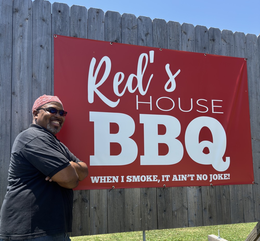 James "Red" Morris stands in front of a sign for his business, Red's House BBQ.