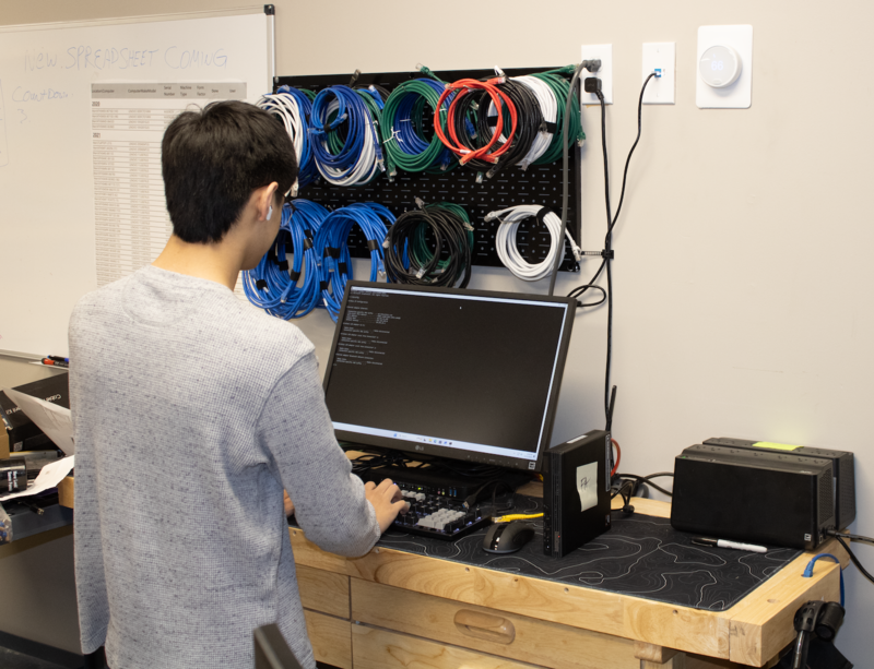 Kaiden Heydt works at a computer workstation, typing on a keyboard while network cables and I.T. equipment are mounted on the wall behind him.