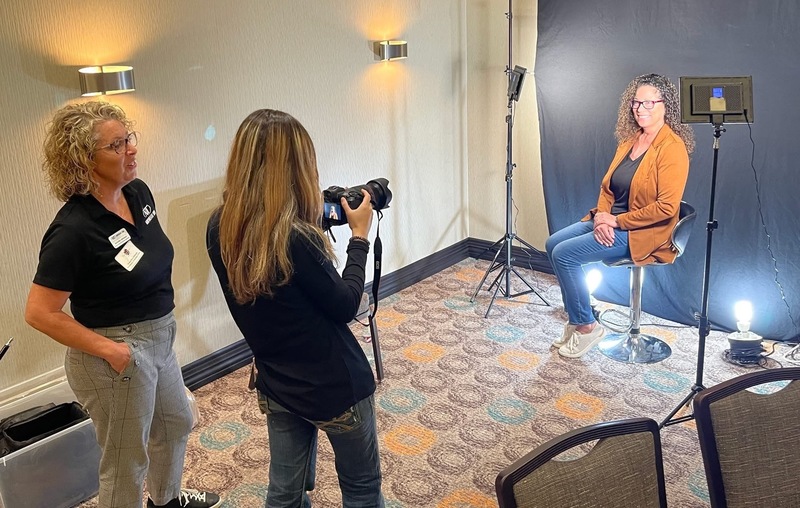 A student photographs a woman seated on a stool during a portrait setup, with studio lights and a backdrop arranged in a conference room.