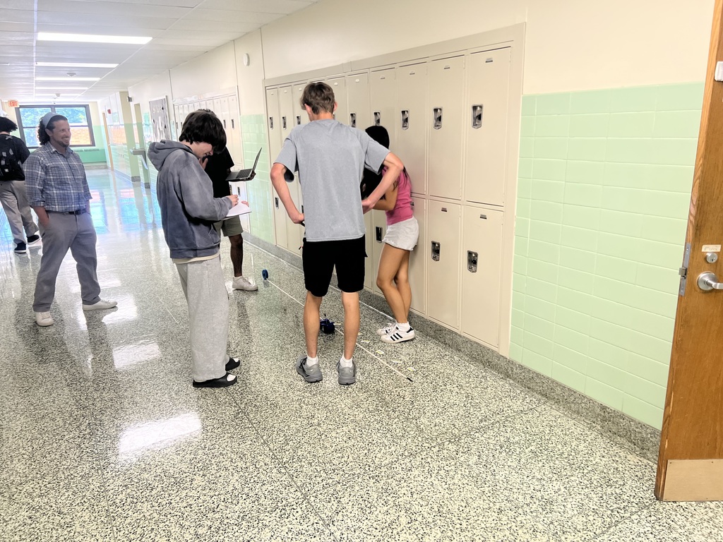 A group of students stands in a school hallway, facing lockers. One student holds papers, another leans on a locker, and some are walking.