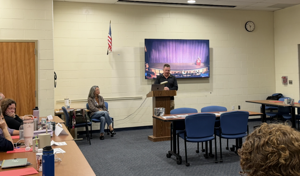 Meeting in a room with a flag and clock on the wall. A man speaks from a podium.