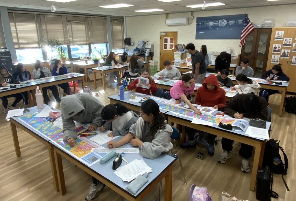Multiple students seated at desks, working on papers and books, inside a classroom with wooden floors.