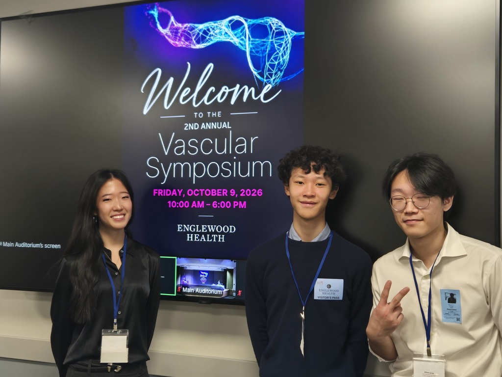 Three people stand next to each other, smiling. A banner behind them says "Welcome to the 2nd Annual Vascular Symposium".