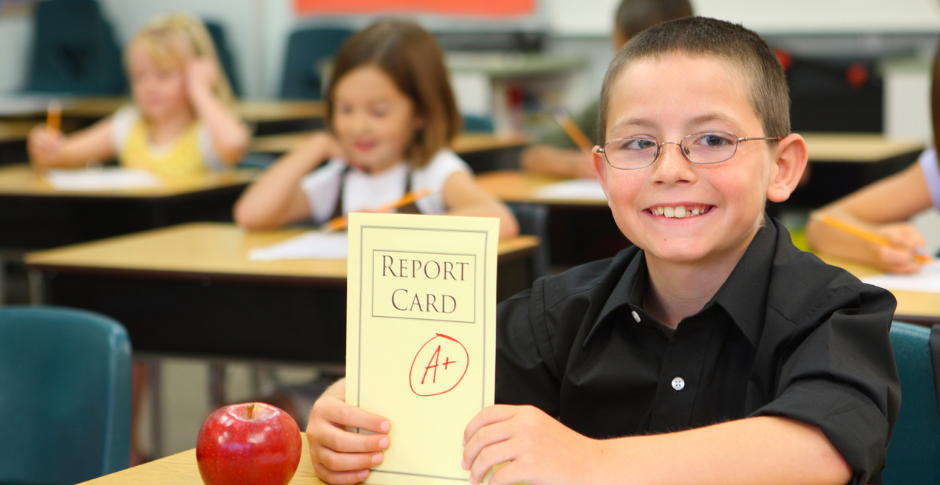 student sitting at desk in classroom holding a report card with A+ on it