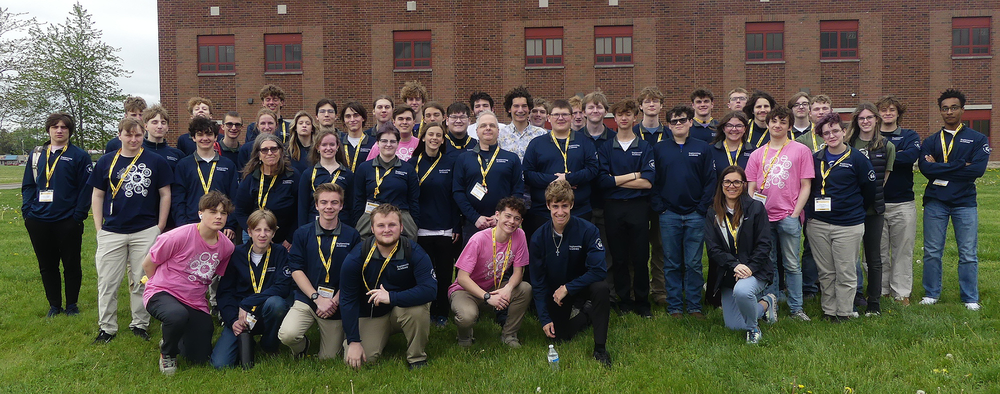 A large group of students on a school trip pose on a lawn in front of a brick building. 