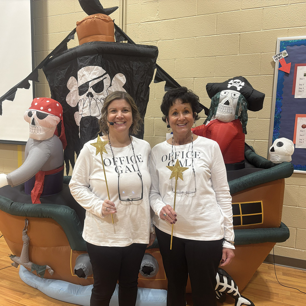 Secretaries from North Ed Transition Campus posing in front of an inflatable pirate ship, wearing matching shirts that say "Office Gal", holding magic wands.